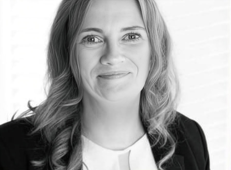 Schwarz-Weiß-Porträt einer lächelnden, blonden Frau im dunklen Blazer und heller Bluse vor hellem Hintergrund.
